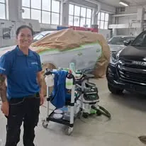 Magdalena står ved arbeidsutstyr i bilverksted, med kjøretøy og reparasjonsarbeid i bakgrunnen.