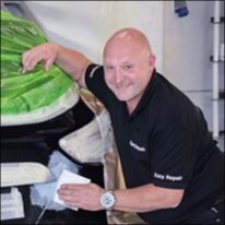 Roy utfører karosserireparasjon på bil med maskerte flater i moderne verkstedmiljø.